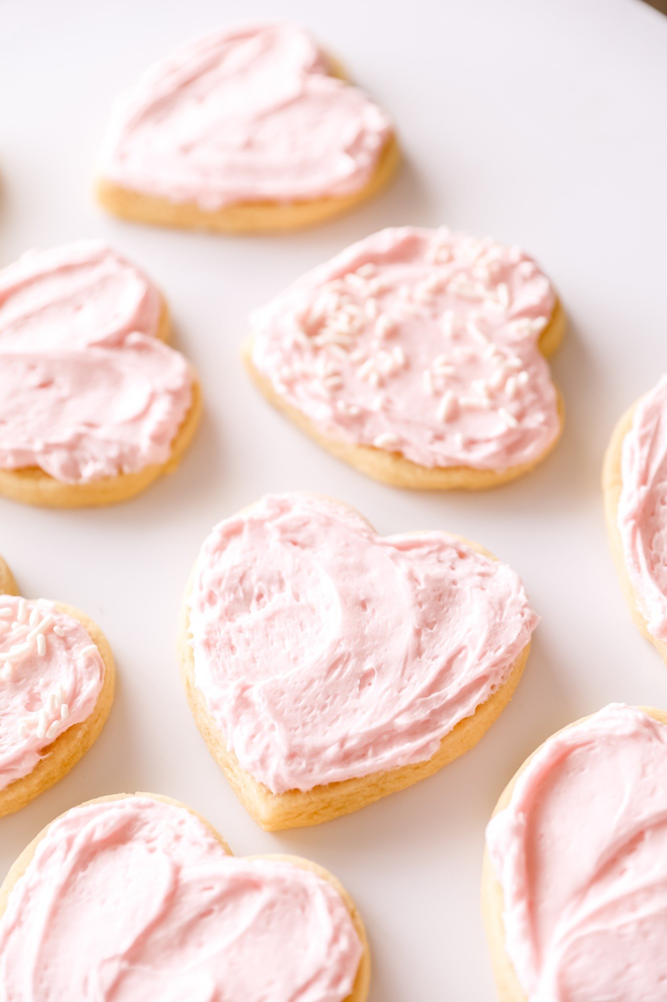 heart-shaped sugar cookies with pink frosting are spread out across a white counter. one cookie has white sprinkles.