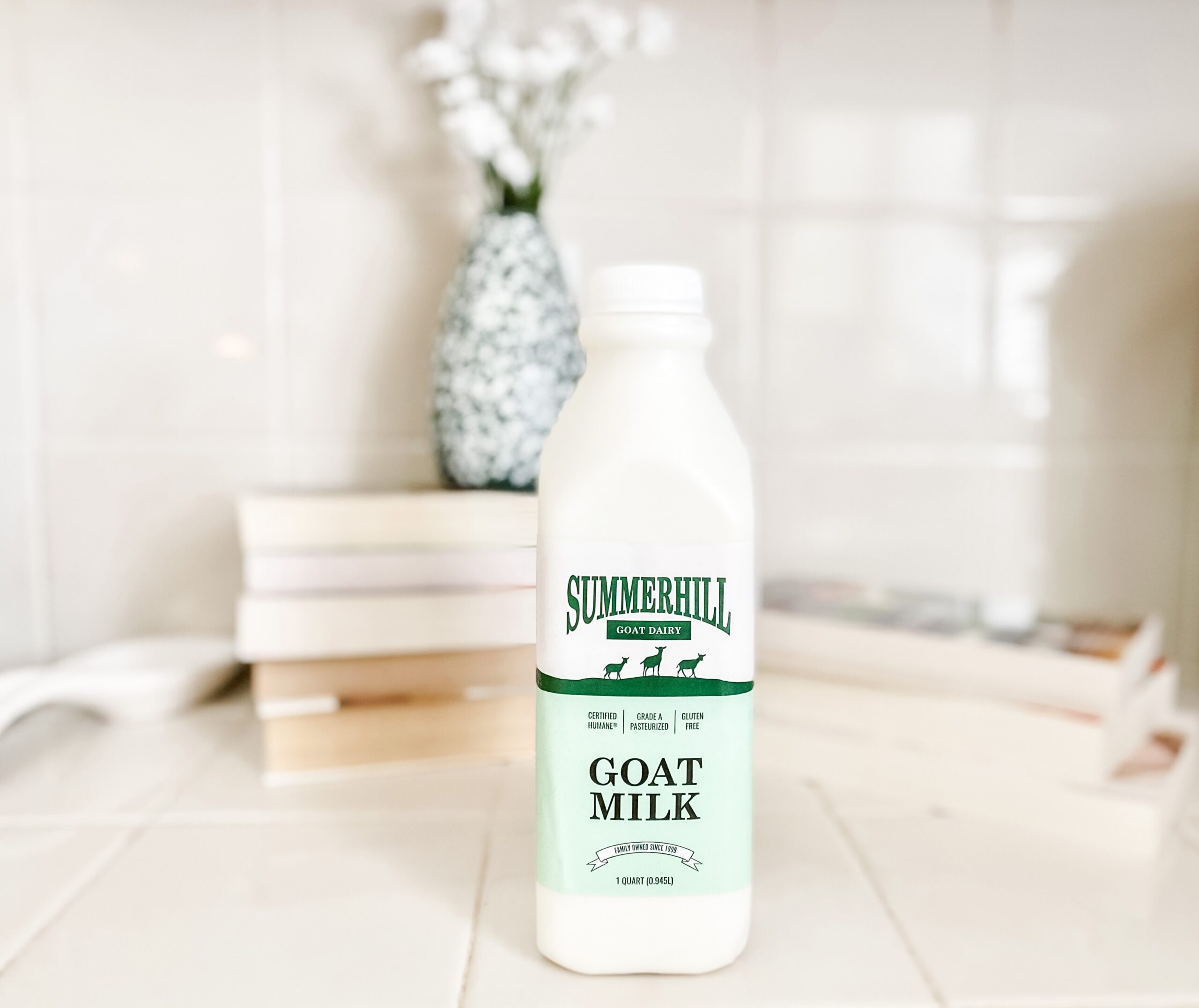 Summerhill goat milk in foreground, with stacks of books, a green vase holding white flowers in the background