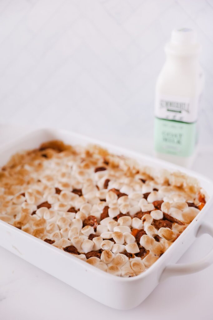 a white casserole dish is filled with sweet potato casserole; orange sweet potatoes are layered with pecans and mini marshmallows. A bottle of Summerhill Goat Milk sits in the right background.