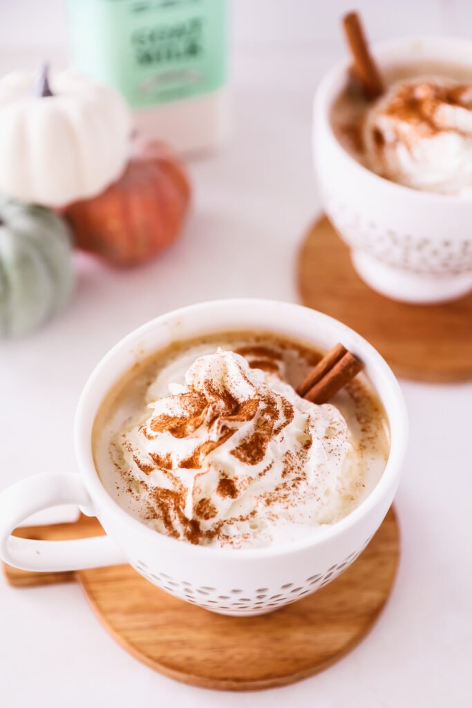 a white teacup full of pumpkin spice latte, whipped cream, and a cinnamon stick sits on top of a small round cutting board. On the left background, there is a blurred out stack of bluegreen, white, and orange pumpkings next to a bottle of Summerhill Goat Milk. A second latte sits in the background on the right.