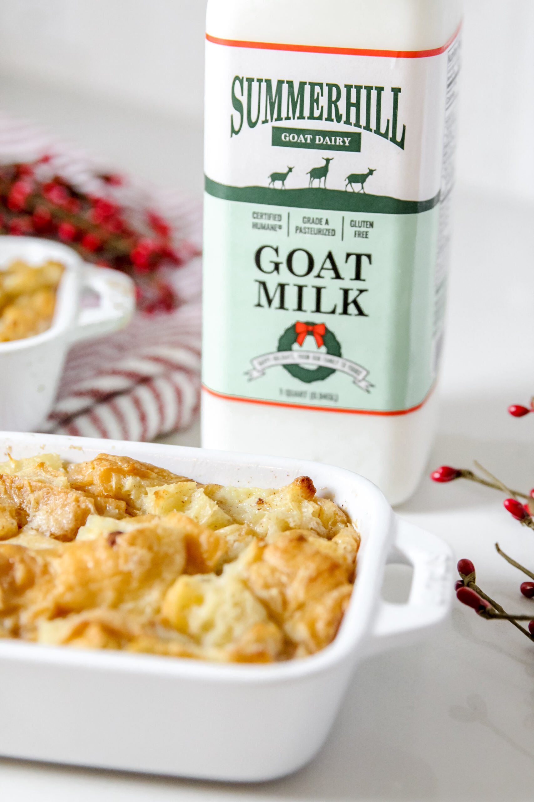 A white Cruset pan of golden brown bread pudding sits in the foreground. The Red and Green version of the Summerhill Goat Milk bottle sits behind it on the right, surrounded by decorative holly berry branches. The corner of a second dish of bread pudding sits in the background on the left on top of a red and white striped towel.