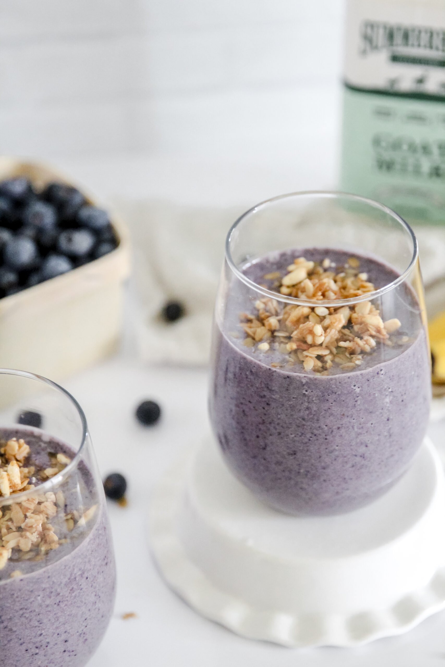 2 glasses of purple blueberry smoothie topped with granola sit in clear glasses. The glass on the right is raised on an upside down white dish. There is a wooden basket of blueberries on the left in the background, and a bottle of Summerhill Goat Milk and two bananas in the background on the right. Granola and blueberries are sprinkled on the white counter for decoration.