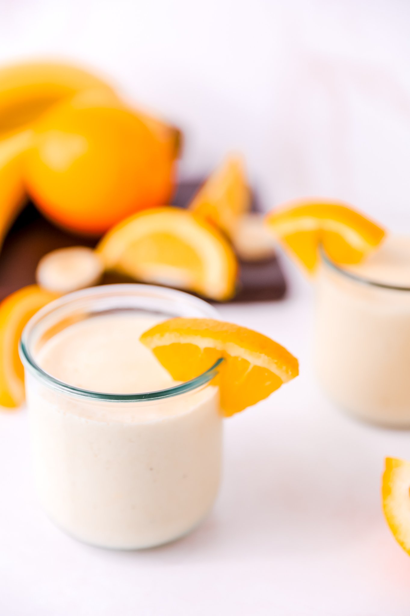 a glass of an orange smoothie with an orange slice garnish sits in the left foreground. There is a second glass in the right background, with a blurred cutting board covered in oranges and orange slices sitting in the left background.