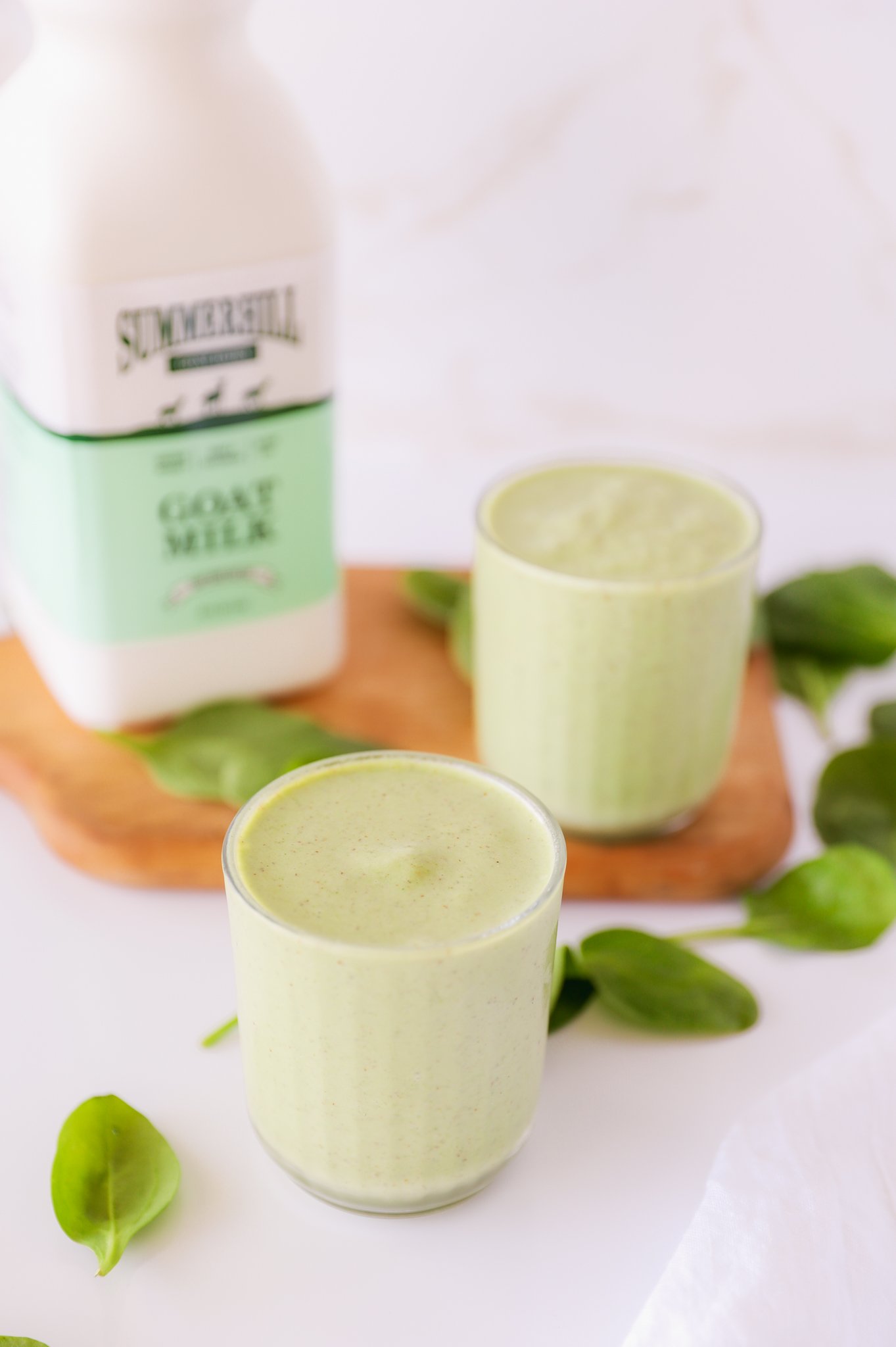 2 glasses of green smoothie, one on the white counter and one on a brown cutting board with a bottle of Summerhill goat milk. Spinach is sprinkled around the counter as a garnish