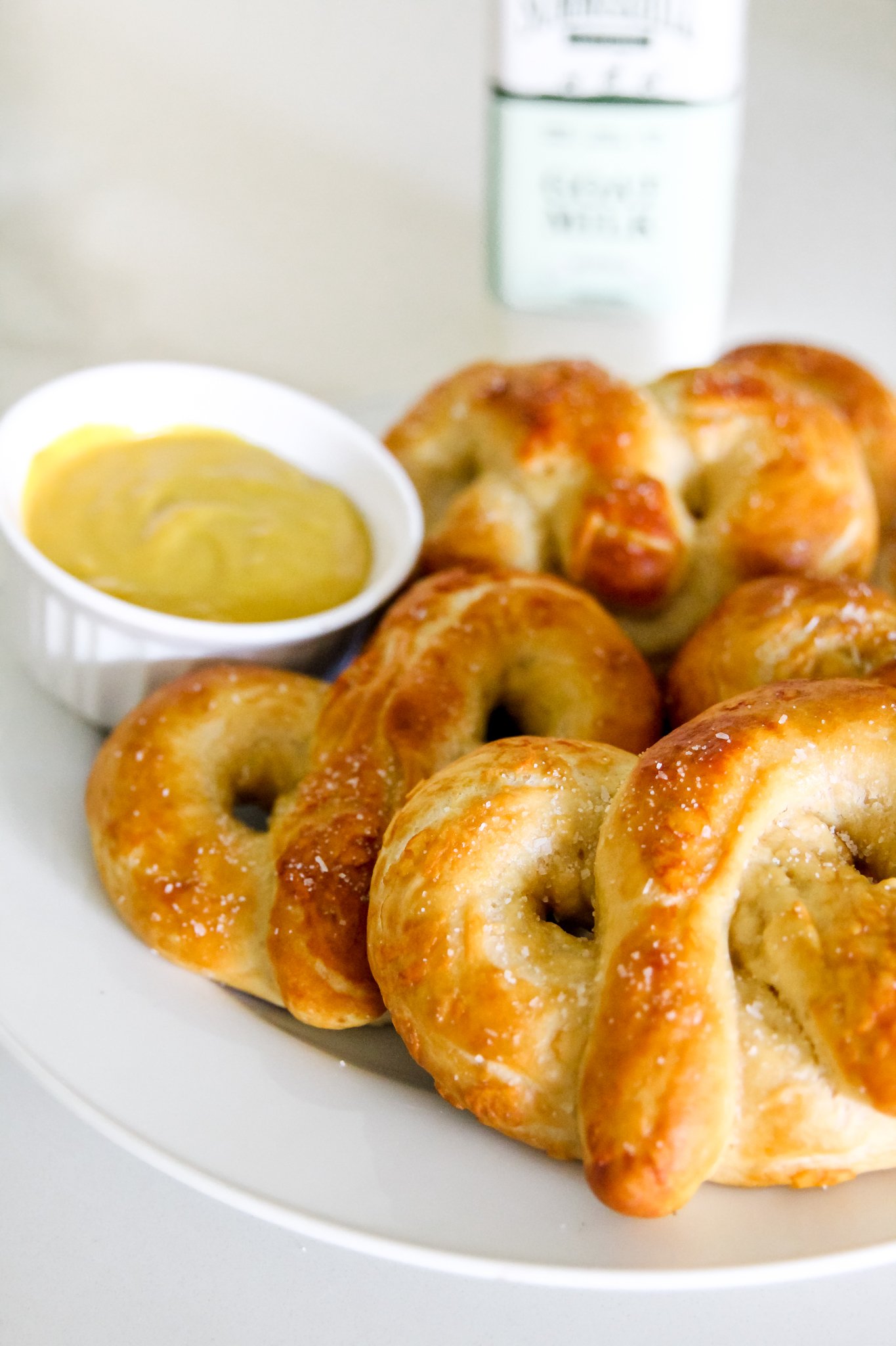 5 pretzels are plated on a white plate, with a yellow sauce in a bowl to the left. A bottle of Summerhill Goat Milk sits blurred in the background on the right.