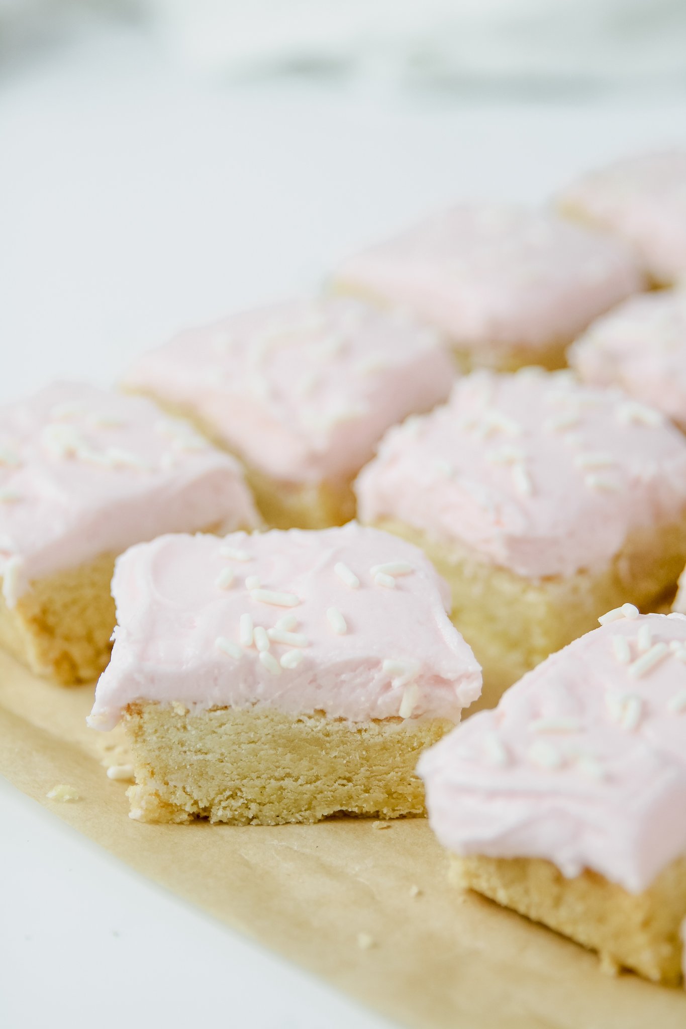 A wooden cutting board on a white counter. diagonal rows of sugar cookie bars with pink frosting and white sprinkles sit diagonally across the photo. 4 bars on the top, 2.5 in the middle, and one on the bottom.