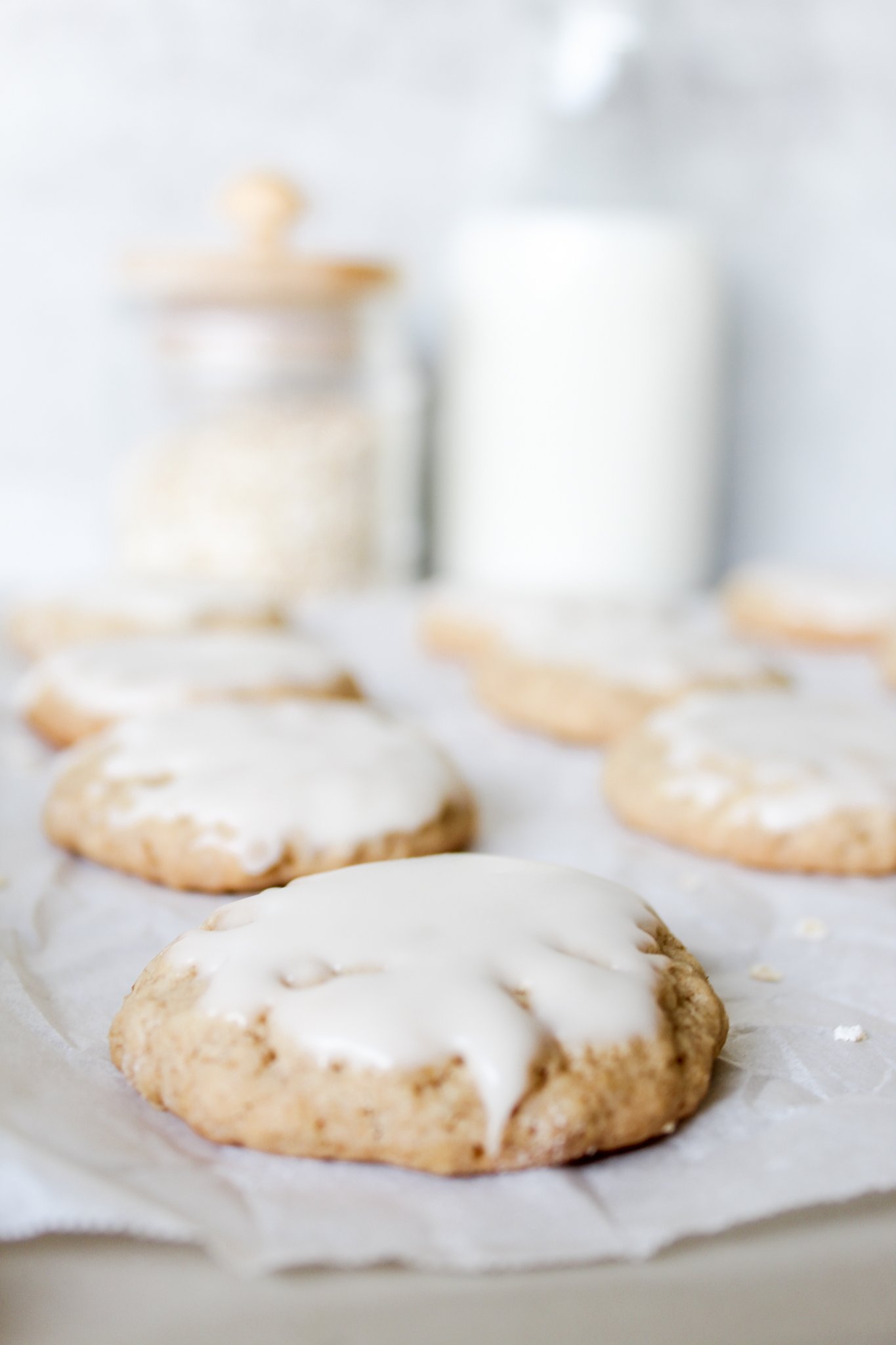 6 oatmeal cookies with white frosting, glass jars of oats and milk in the background