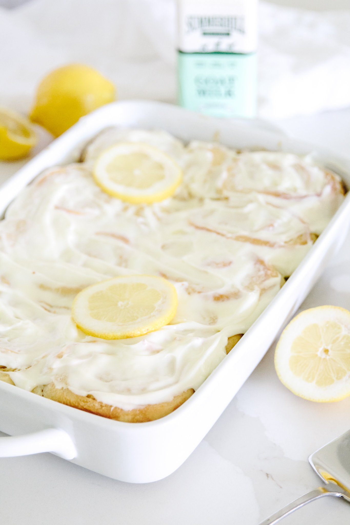 A white casserole dish full of frosted lemon rolls sits on a white counter. There are two circular lemon slices on the rolls for garnish. There is a lemon half on the right foreground, and a lemon and a half lemon and Summerhill goat milk in the background.
