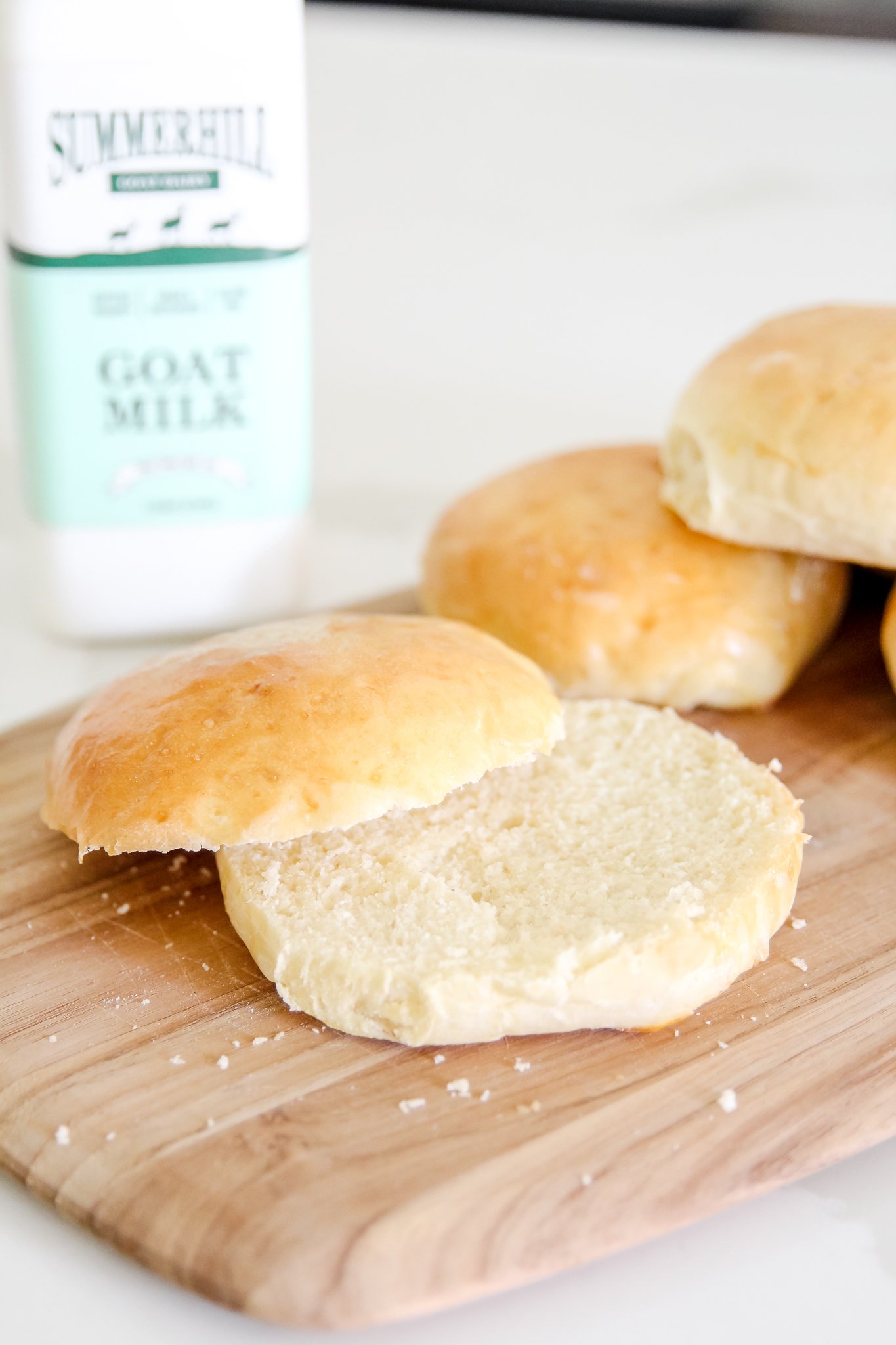 a sliced hamburger bun and 2 unsliced hamburger buns sit on a wooden cutting board. a blurred bottle of Summerhill Goat Milk sits in the left background.