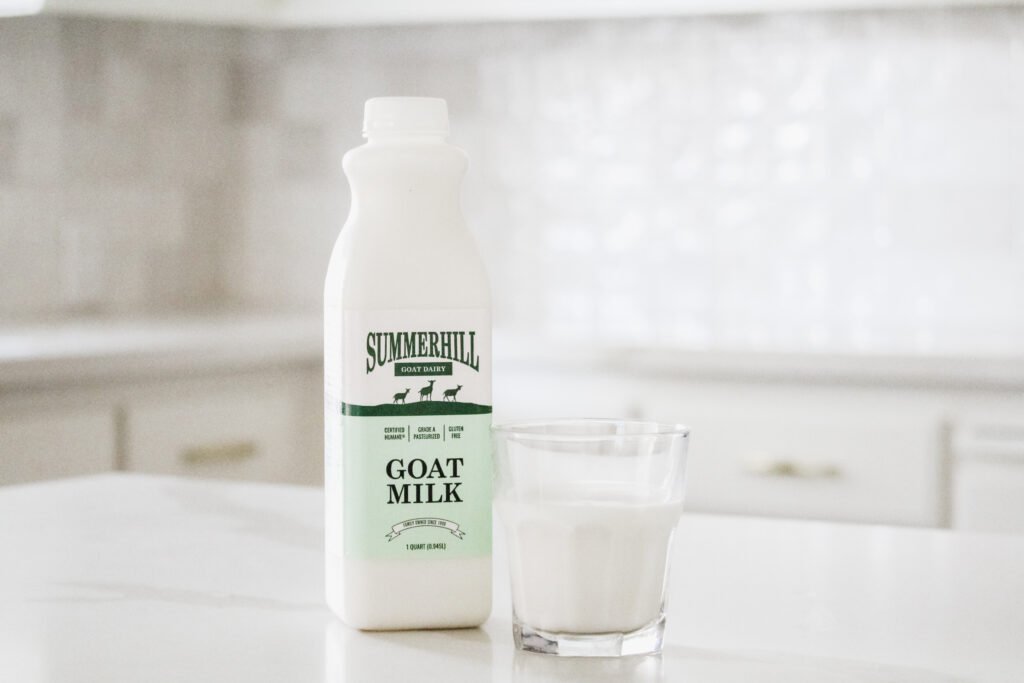 A white kitchen. A bottle of Summerhill Goat Milk sits Center Left on the white counter, and a 8 oz glass of goat milk sits Center Right.