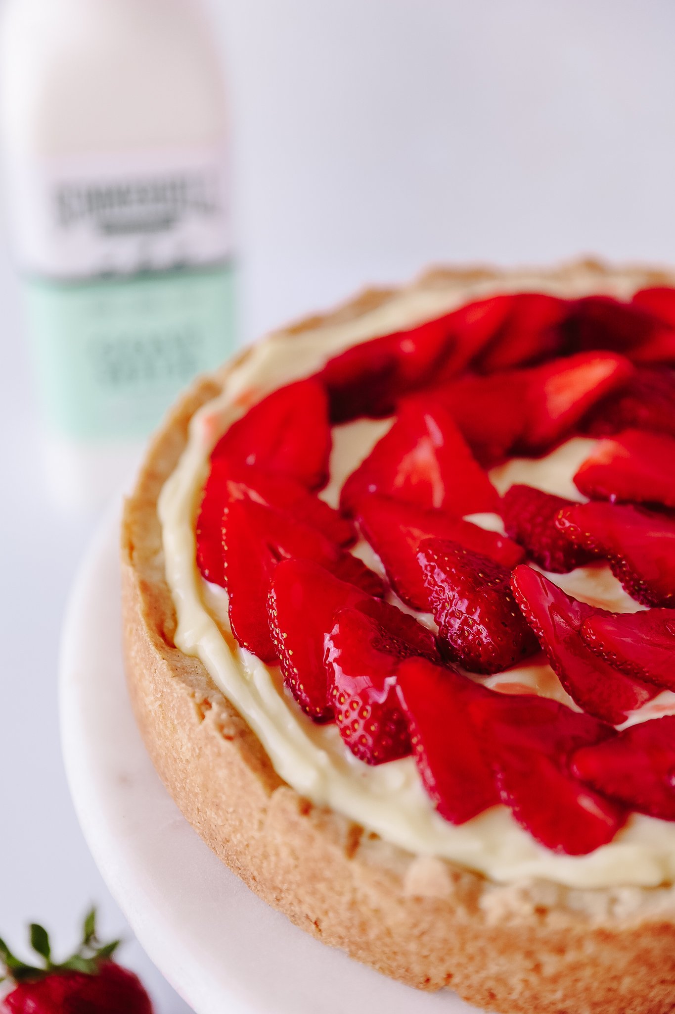 Photo is of an 8inch tart with custard cream and sliced strawberries. A blurred bottle of Summerhill Goat Milk is in the back left of the photo.