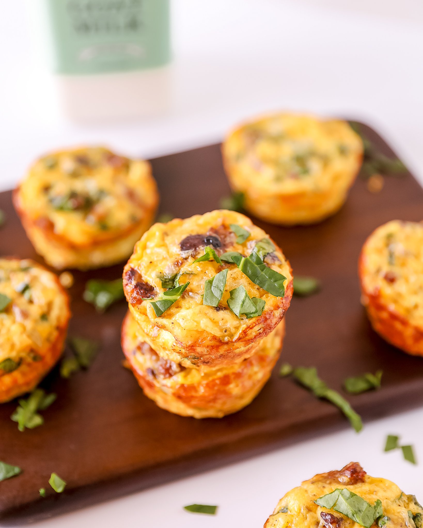 2 golden brown egg bites are stacked in focus on a dark wooden cutting board, surrounded by blurred golden egg bites. A bottle of Summerhill goat milk is blurred in the left background. the food is covered in chopped basil for garnish.