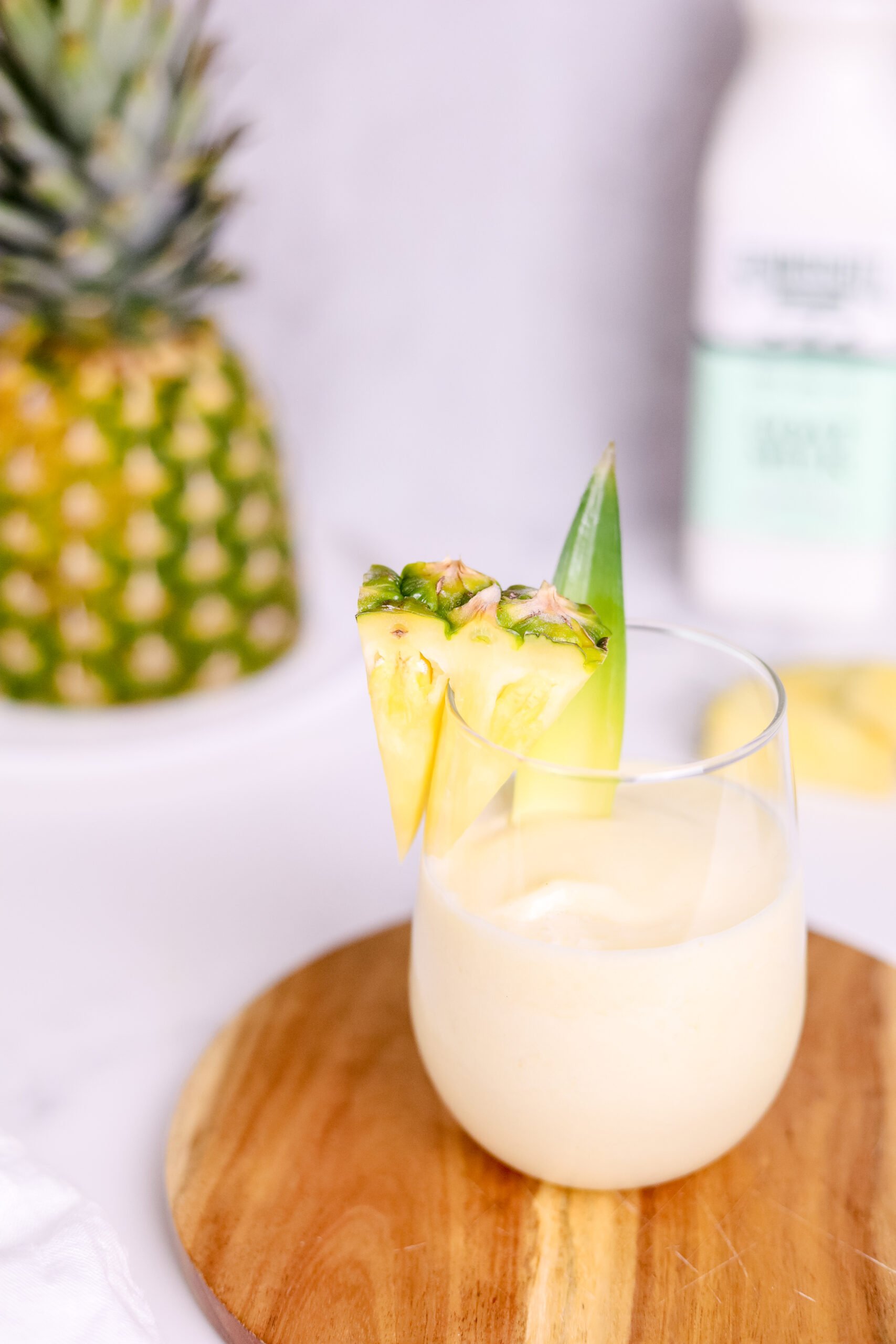 In the foreground: a small round wooden cutting board sits underneath a stemless wine glass filled with the pina colada and garnished with a triangular slice of pineapple. In the background, the top half of the pineapple sits on the left, and a blurred out bottle of Summerhill Goat Milk on the right.