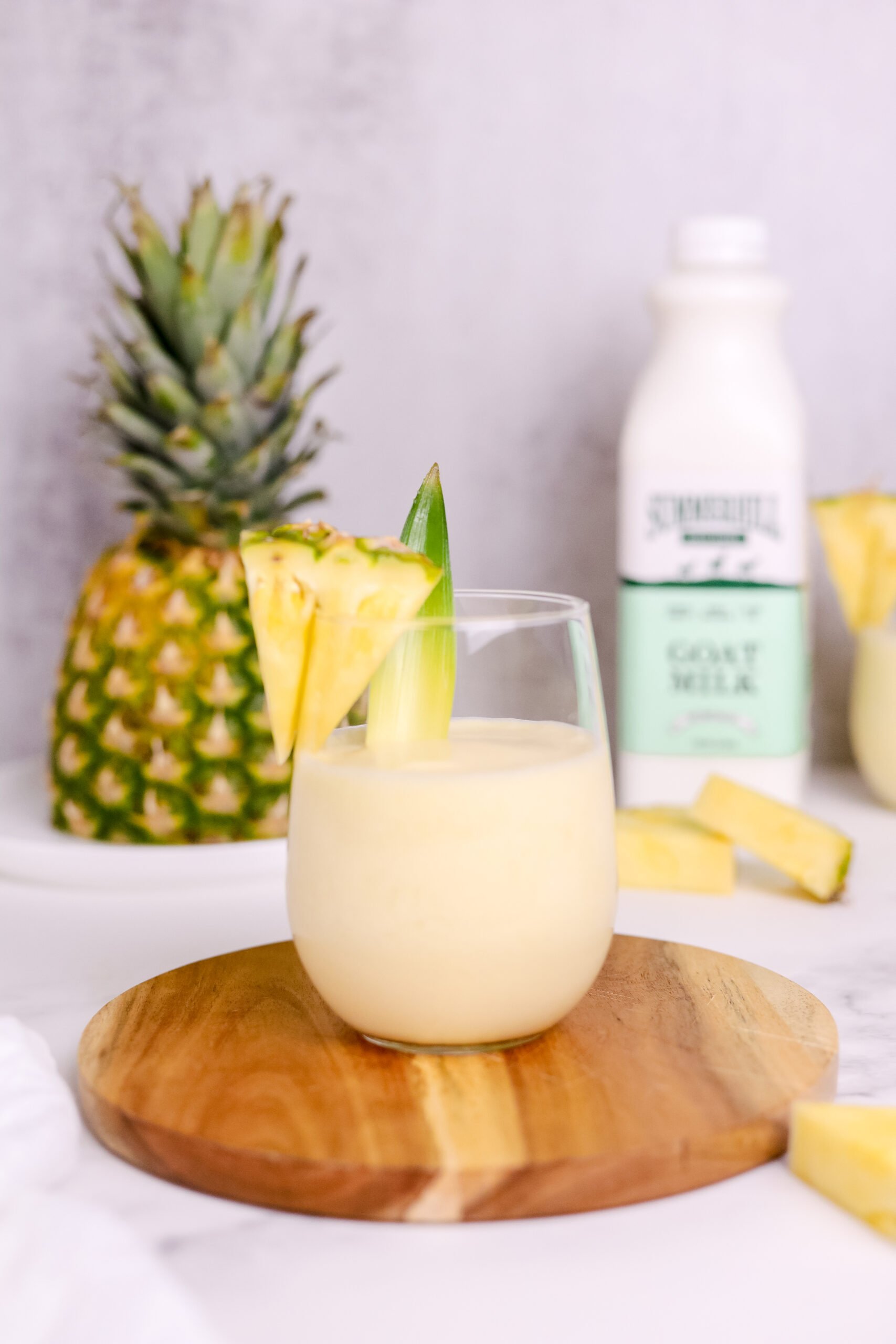 In the foreground: a small round wooden cutting board sits underneath a stemless wine glass filled with the pina colada and garnished with a triangular slice of pineapple. In the background, the top half of the pineapple sits on the left, and a blurred out bottle of Summerhill Goat Milk on the right.