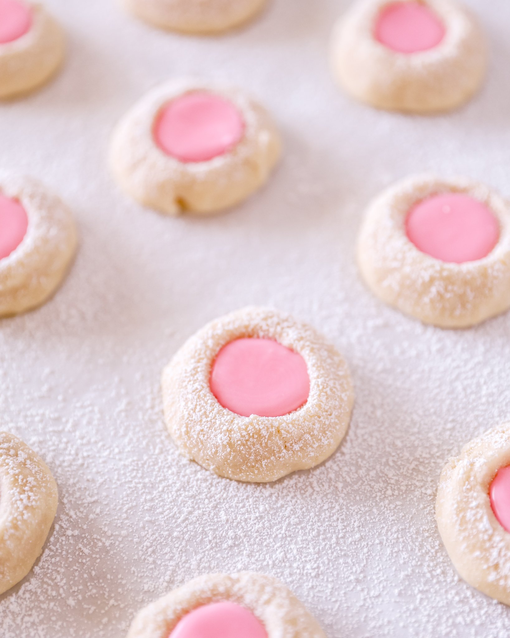 thumprint cookies with a pink frosting center sit on a baking sheet covered in powdered sugar.