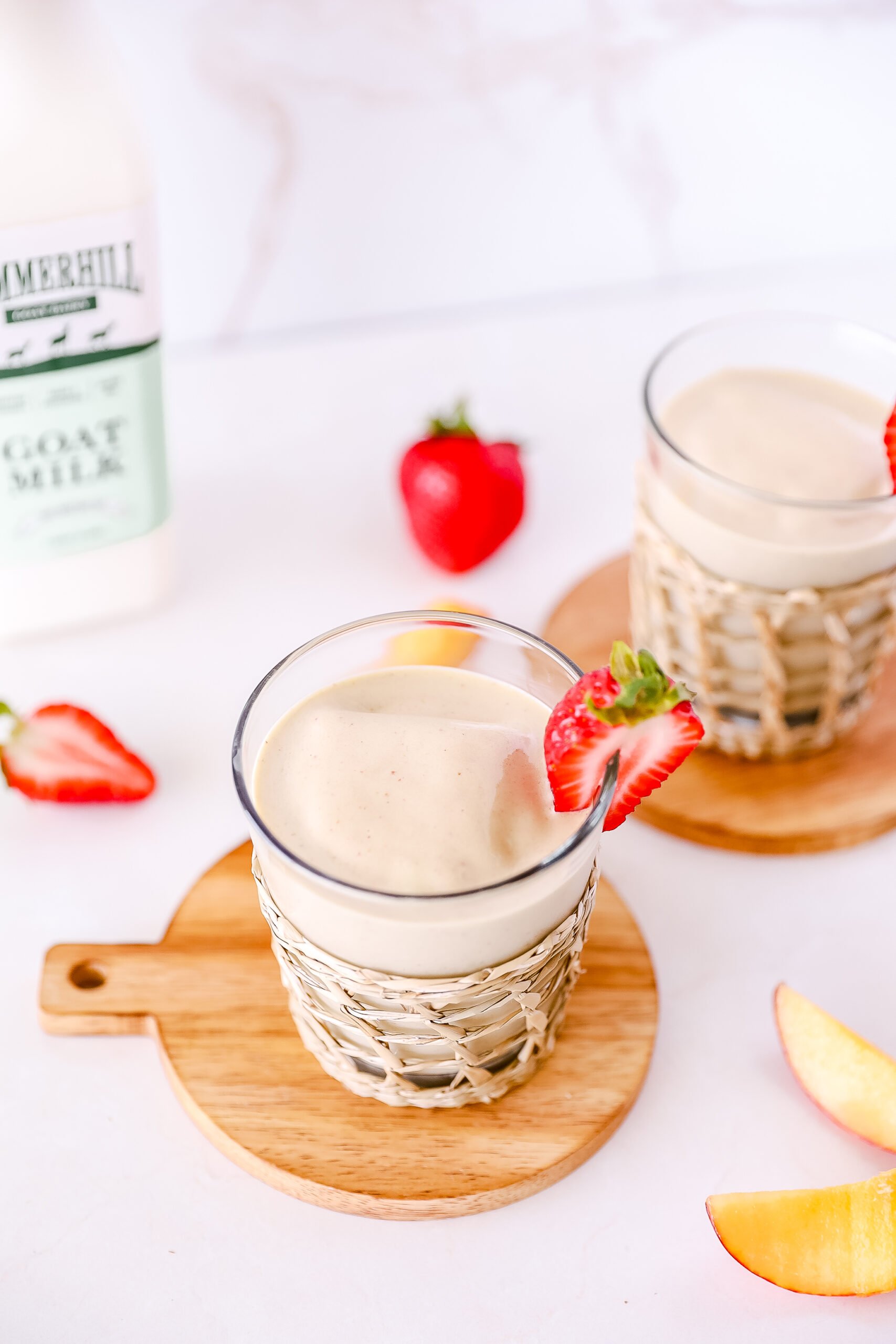 photo taken from angle above. A glass of peach smoothie sits on a small round cutting board, with a wicker wrap and strawberry slice for garnish. A bottle of Summerhill goat milk sits in the left background. peach slices and strawberries are scattered on the counter for decoration.
