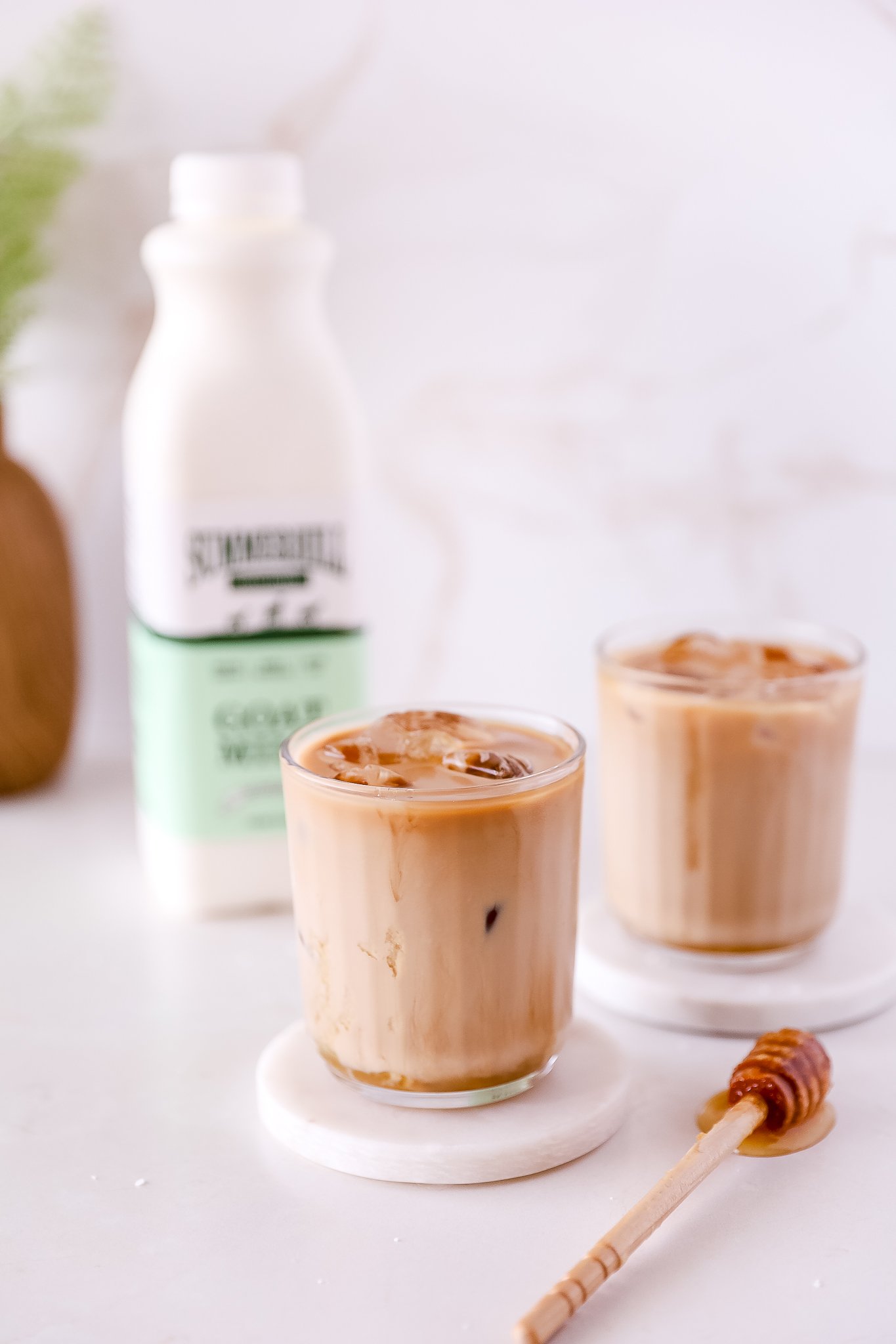A glass of salted honey latte sits on a white coaster in the foreground, with a honey wand on the counter next to it. Behind it there is a bottle of Summerhill Goat Milk on the left and another latte glass on the right. A potted plant is blurrred into the left corner.