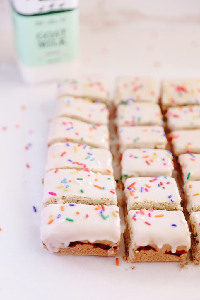 2 columns of small rectangular pop tarts. They have a strawberry filling and rainbow sprinkles. There is a bottle of Summerhill Goat Milk in the top left corner in the background