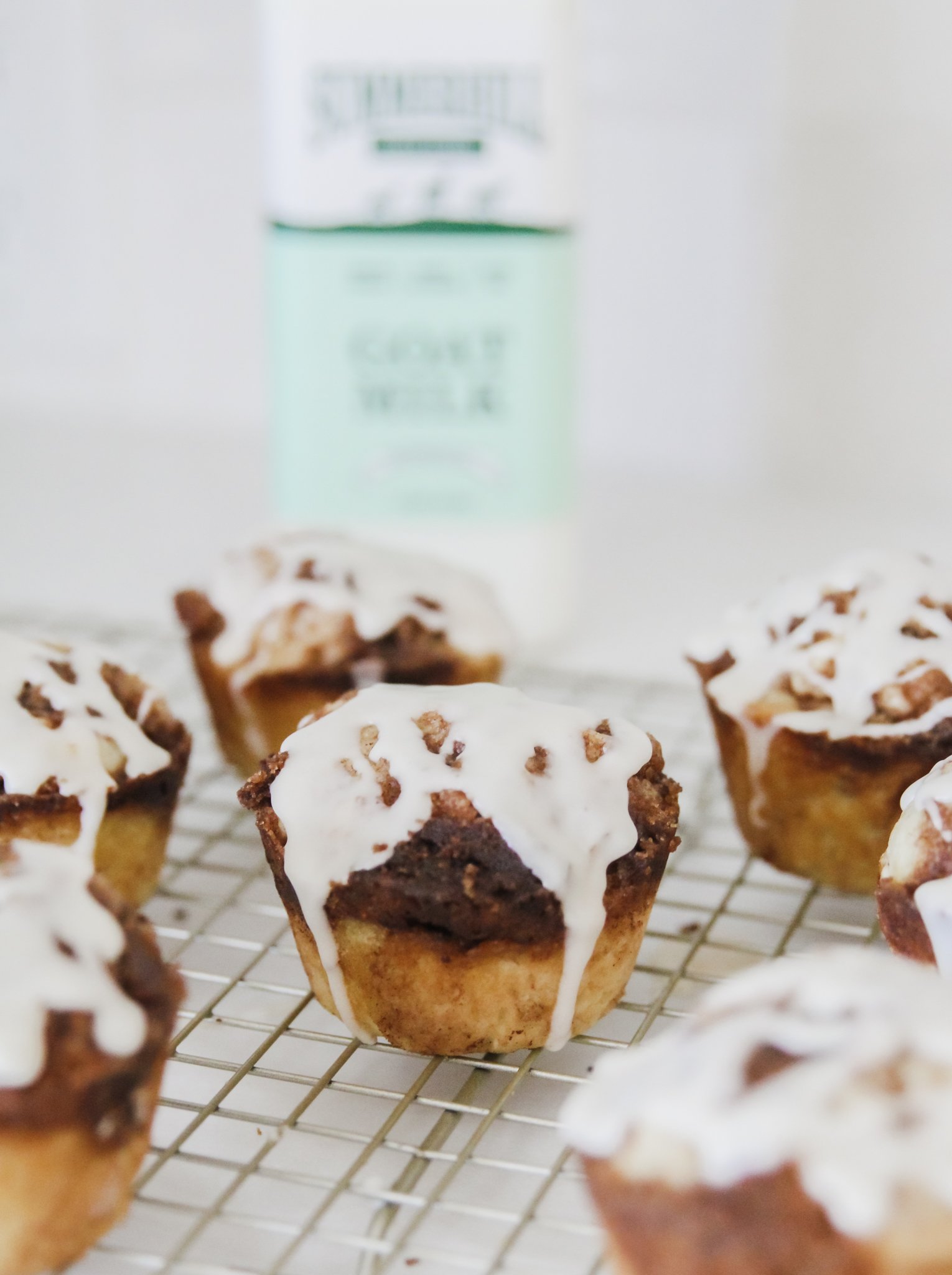 7 cinnamon roll muffins with drizzled icing sit on top of a wire cooling rack, with a blurred bottle of Summerhill Goat Milk in the background.
