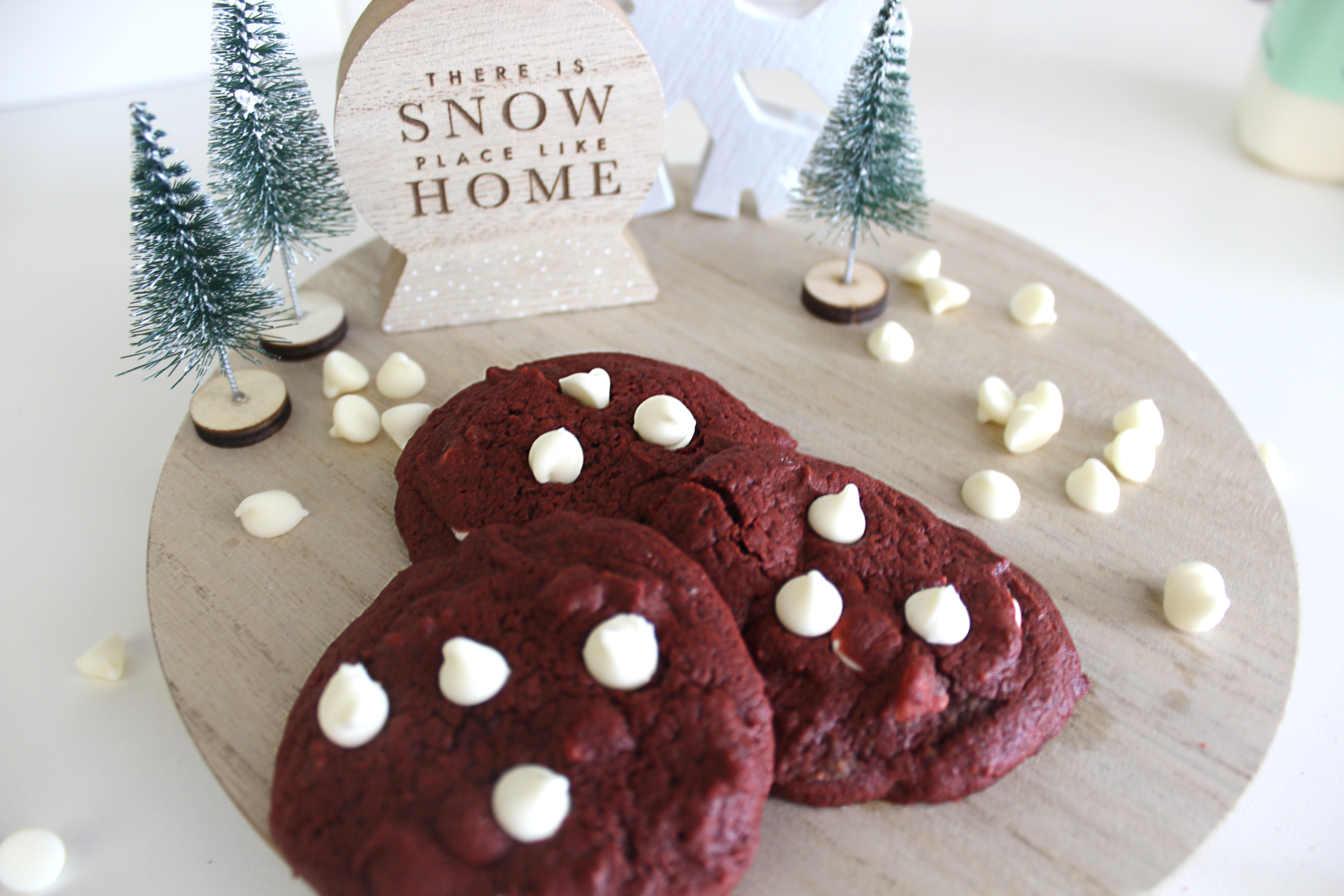 wooden circle tray on white counter. 3 red velvet cookies with white chocolate chips sit in foreground. tray is decorated with white chocolate chips sprinled throughout and in background, a small wooden snowglobe-shaped figurine says "There's SNOW place like HOME" surrounded by 3 Christmas trees.